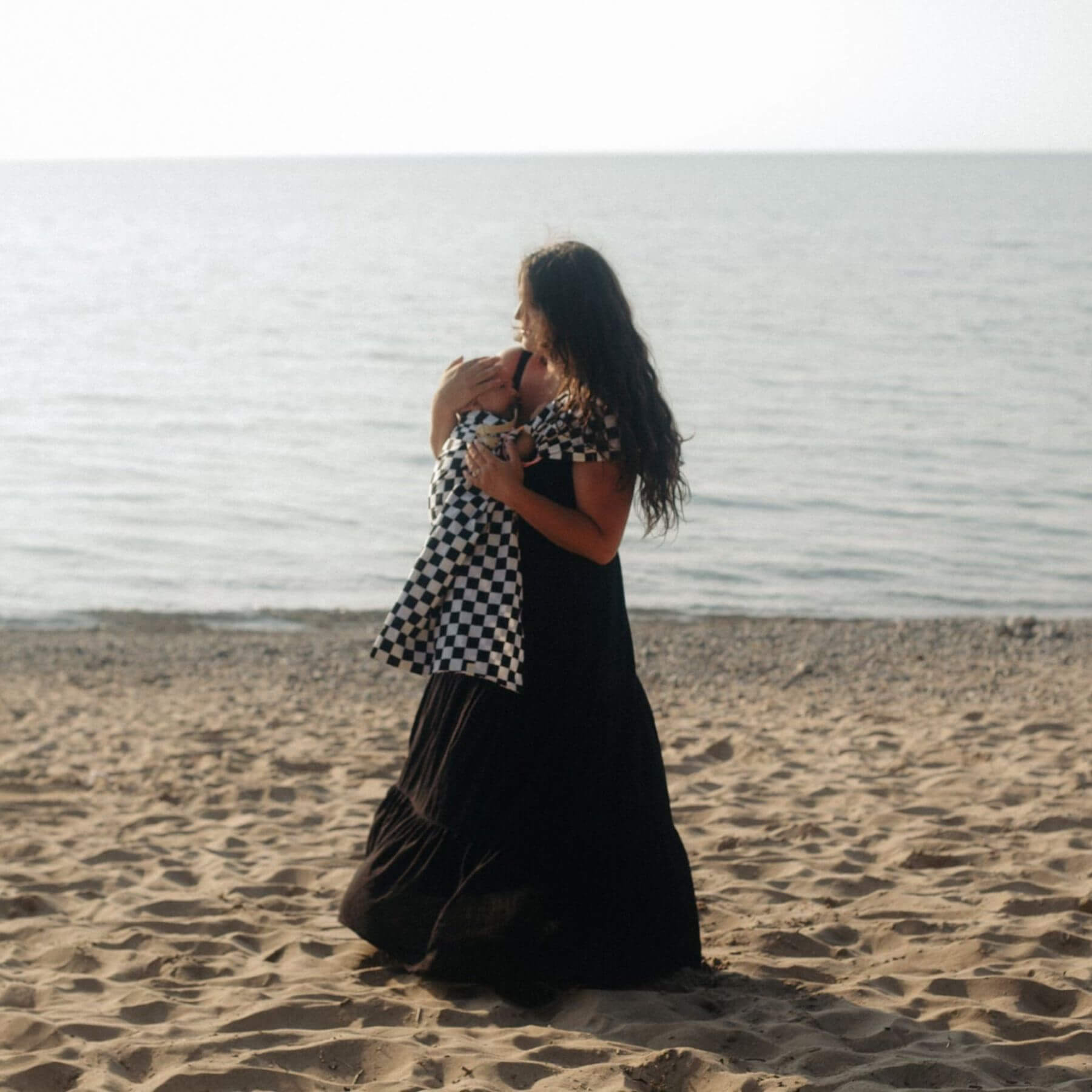 Woman in a black dress holding a baby in Little Mae & Co Race Ring Sling Carrier on a sandy beach.