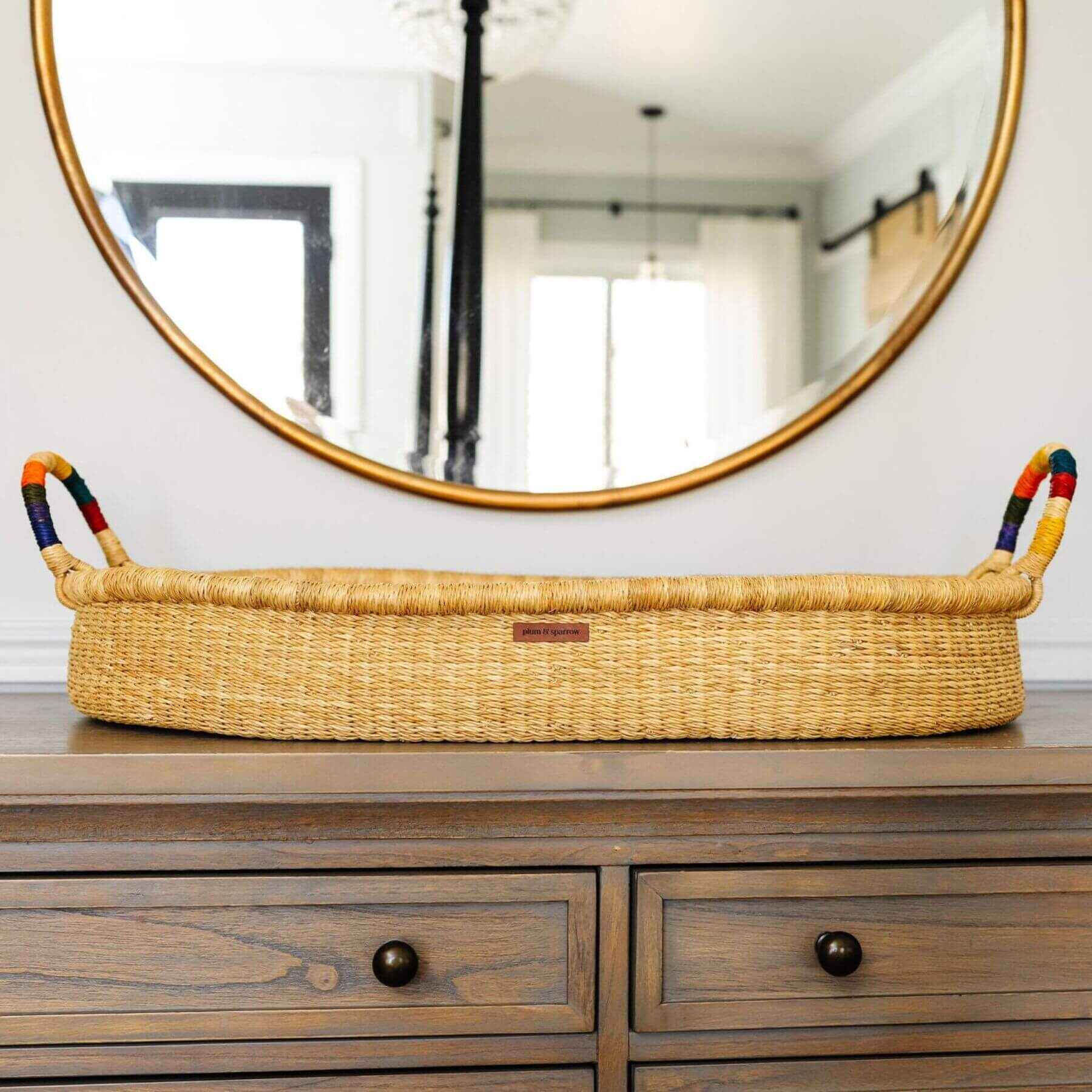 Plum+Sparrow Rainbow Handle Changing Basket on a wooden dresser below a round mirror.