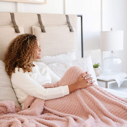 Woman lying on a bed with Ribbed Bamboni® Throw Blankets Cameo and pillows in a bright bedroom.