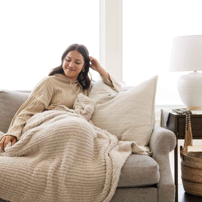 Woman sitting on a couch with Ribbed Bamboni® Throw Blankets Moonbeam and pillow in a cozy living room.