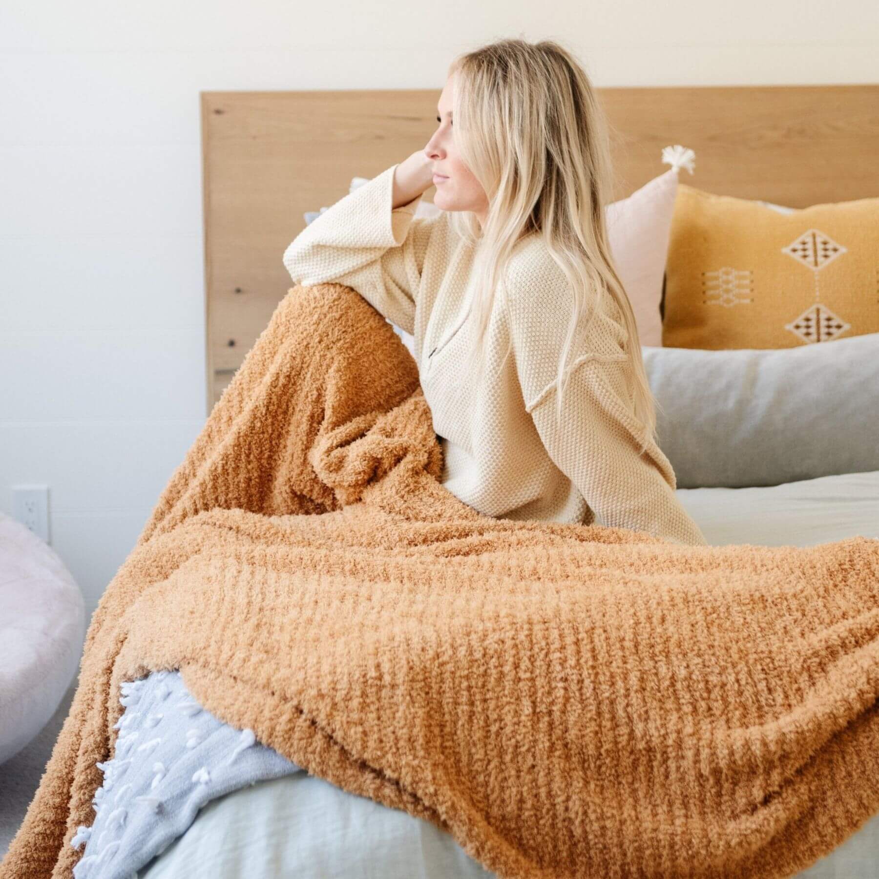 Woman sitting on a bed with Saranoni Ribbed Bamboni® XL Blanket - Golden, surrounded by pillows.