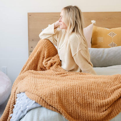 Woman sitting on a bed with Saranoni Ribbed Bamboni® XL Blanket - Golden, surrounded by pillows.