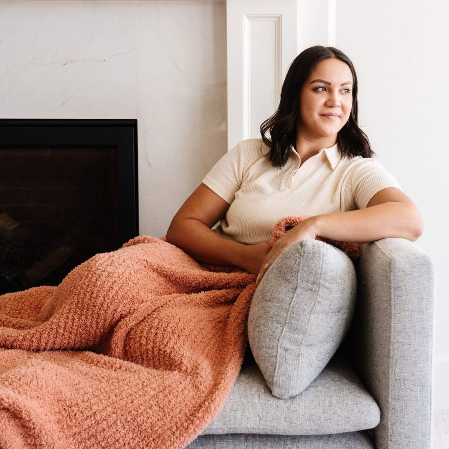 Woman sitting on a couch with Saranoni Ribbed Bamboni® XL Blanket - Sun-Kissed and gray pillow, smiling.