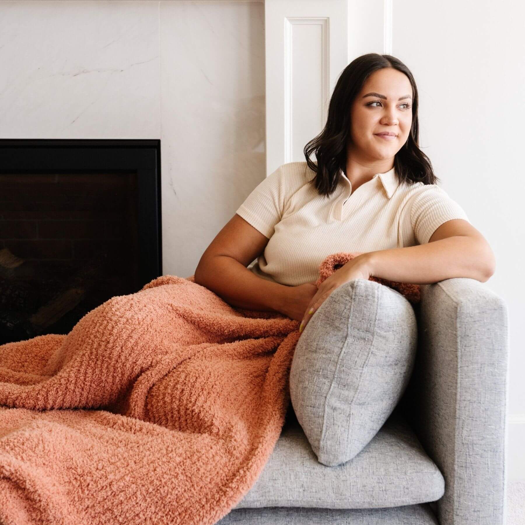 Woman sitting on a couch with Saranoni Ribbed Bamboni® XL Blanket - Sun-Kissed and gray pillow, smiling.