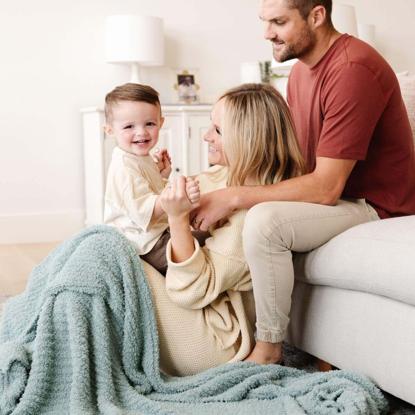 Family of three sitting on a couch with a baby being held by the mother, all smiling.