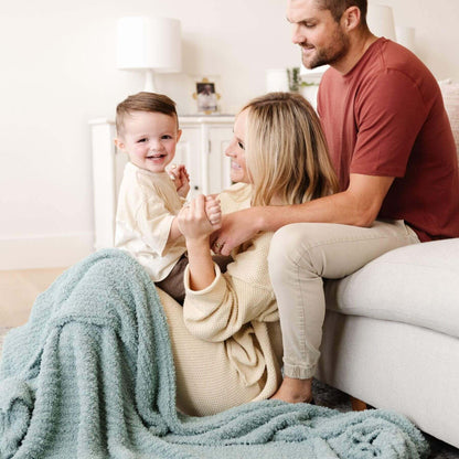 Family of three sitting on a couch with a baby being held by the mother, all smiling.