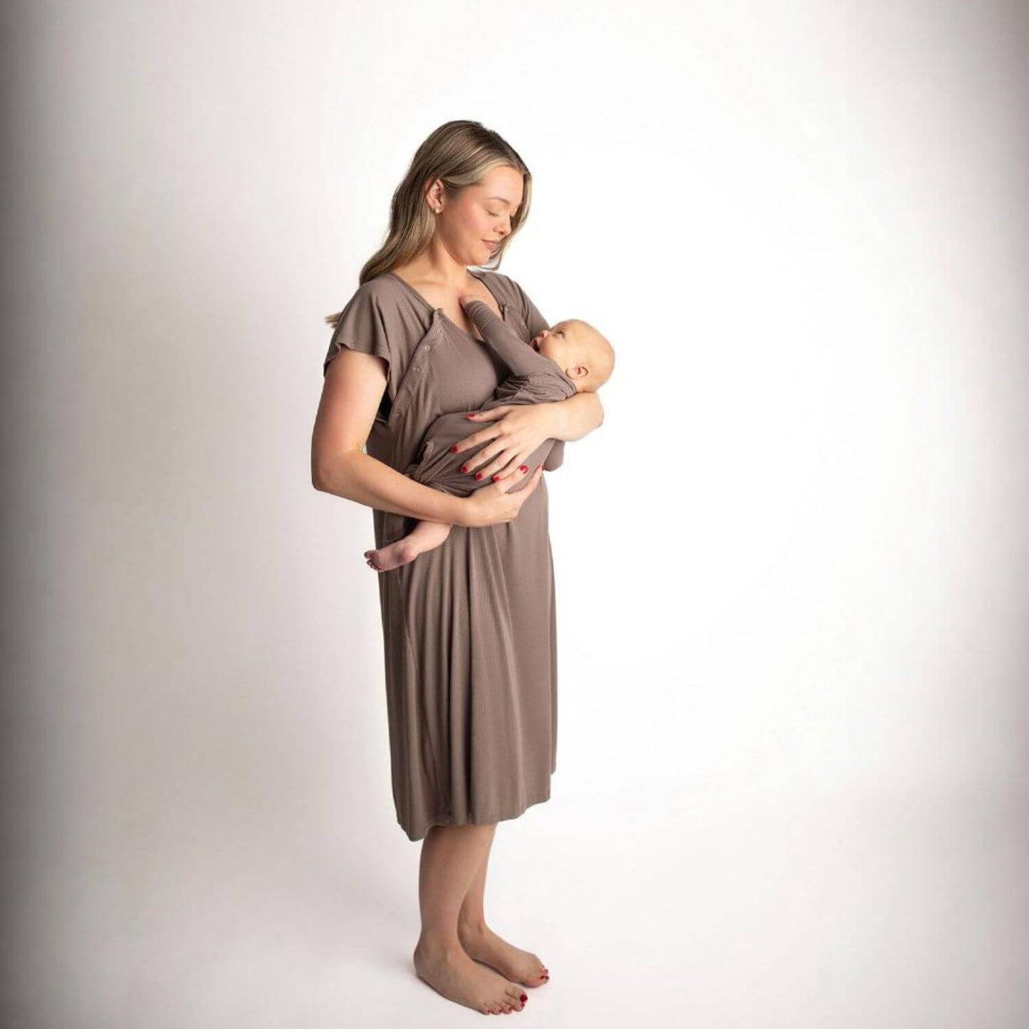 A woman wearing Three Little Tots Ribbed Cocoa Brown Mommy Gown holds a sleeping baby against her chest in a studio setting with a white background.