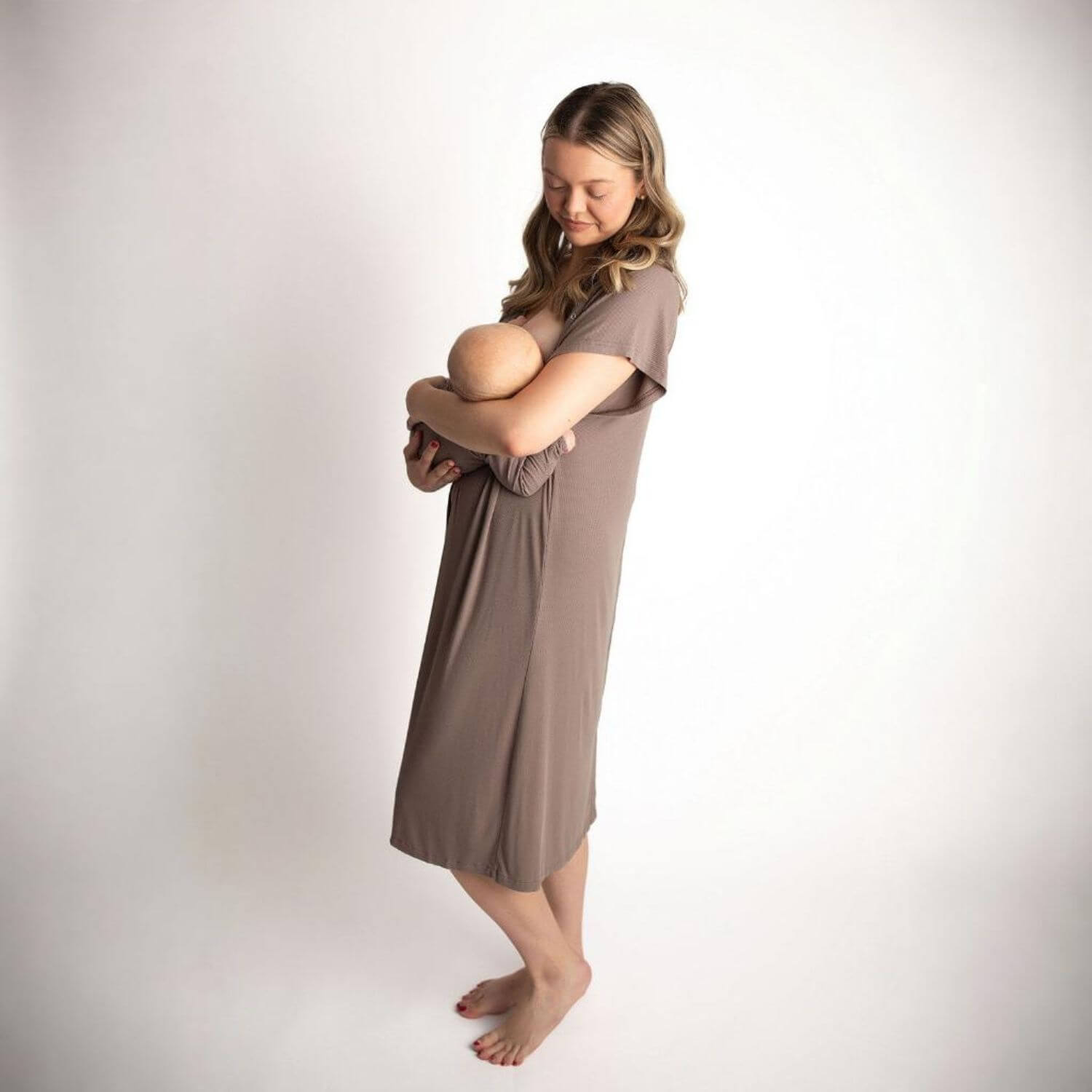 A woman wearing Three Little Tots Ribbed Cocoa Brown Mommy Gown holds and breastfeeds a baby against a plain white studio background.