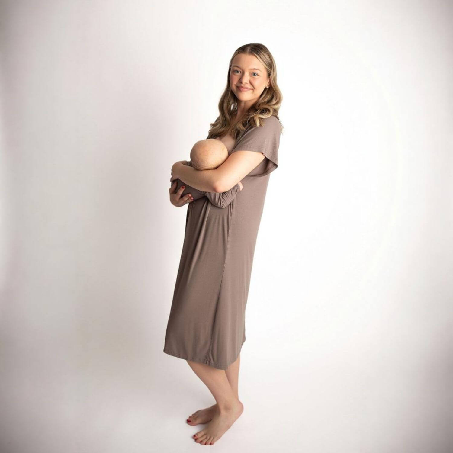 A pregnant woman wearing Three Little Tots Ribbed Cocoa Brown Mommy Gown, holding a baby against her chest while standing against a plain white studio background.