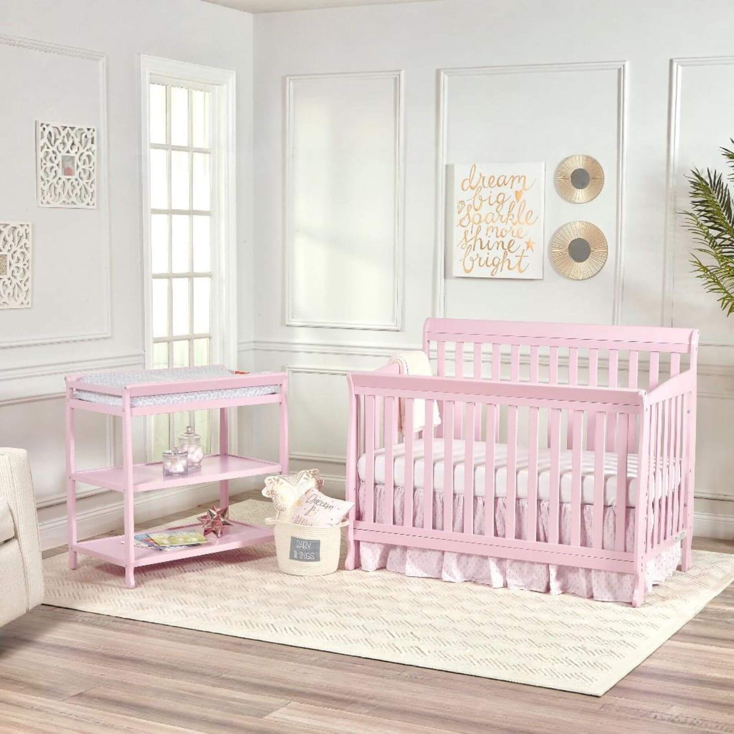 Pink crib and changing table in a nursery with white walls and decor.