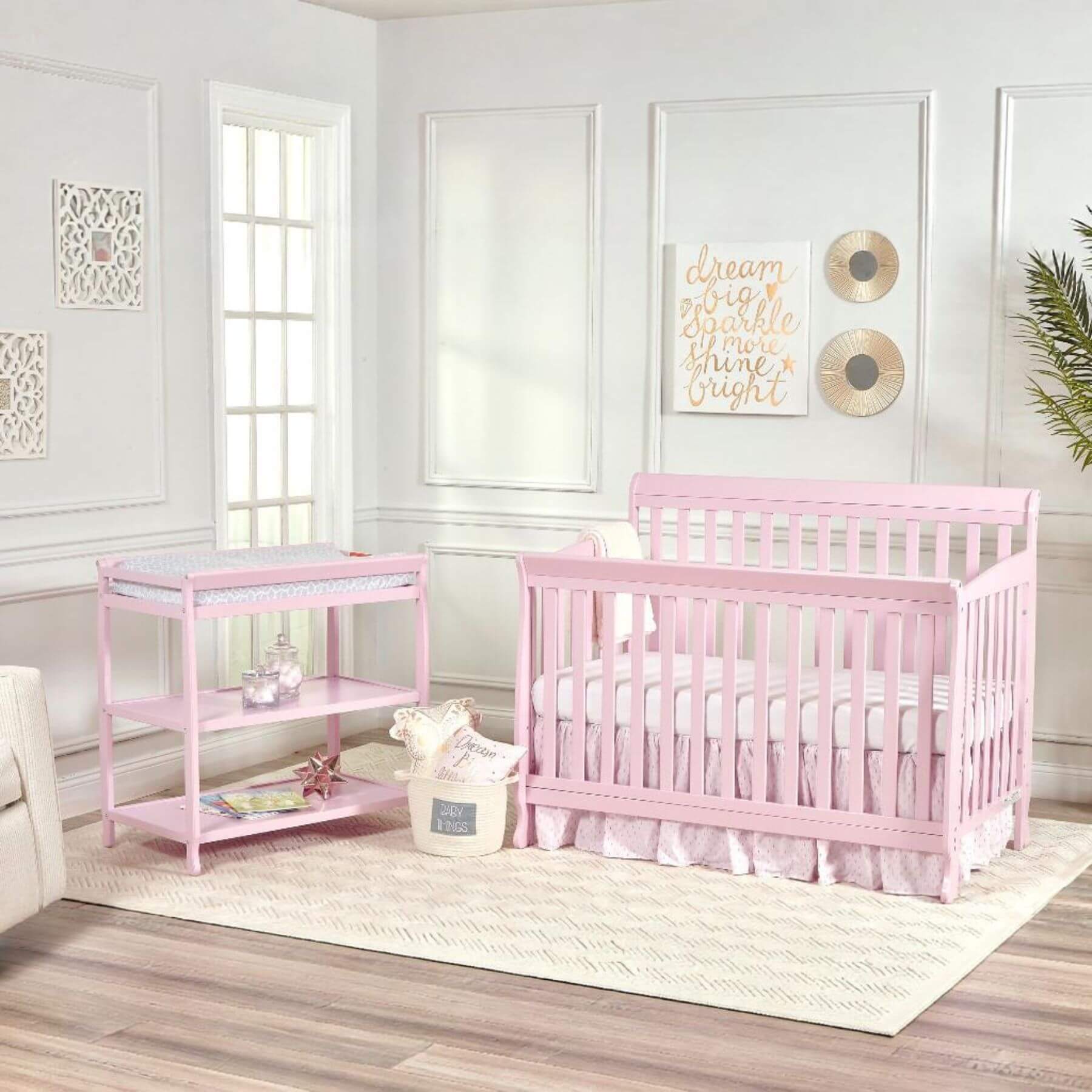 Pink crib and changing table in a nursery with white walls and decor.