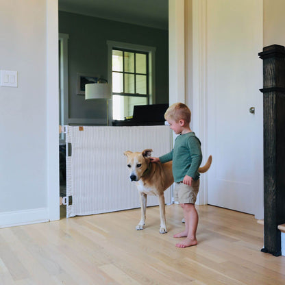 A young boy in a green shirt and khaki shorts stands barefoot on a light wood floor, petting a tan and white dog next to a white ribbed pet gate in a bright home interior.