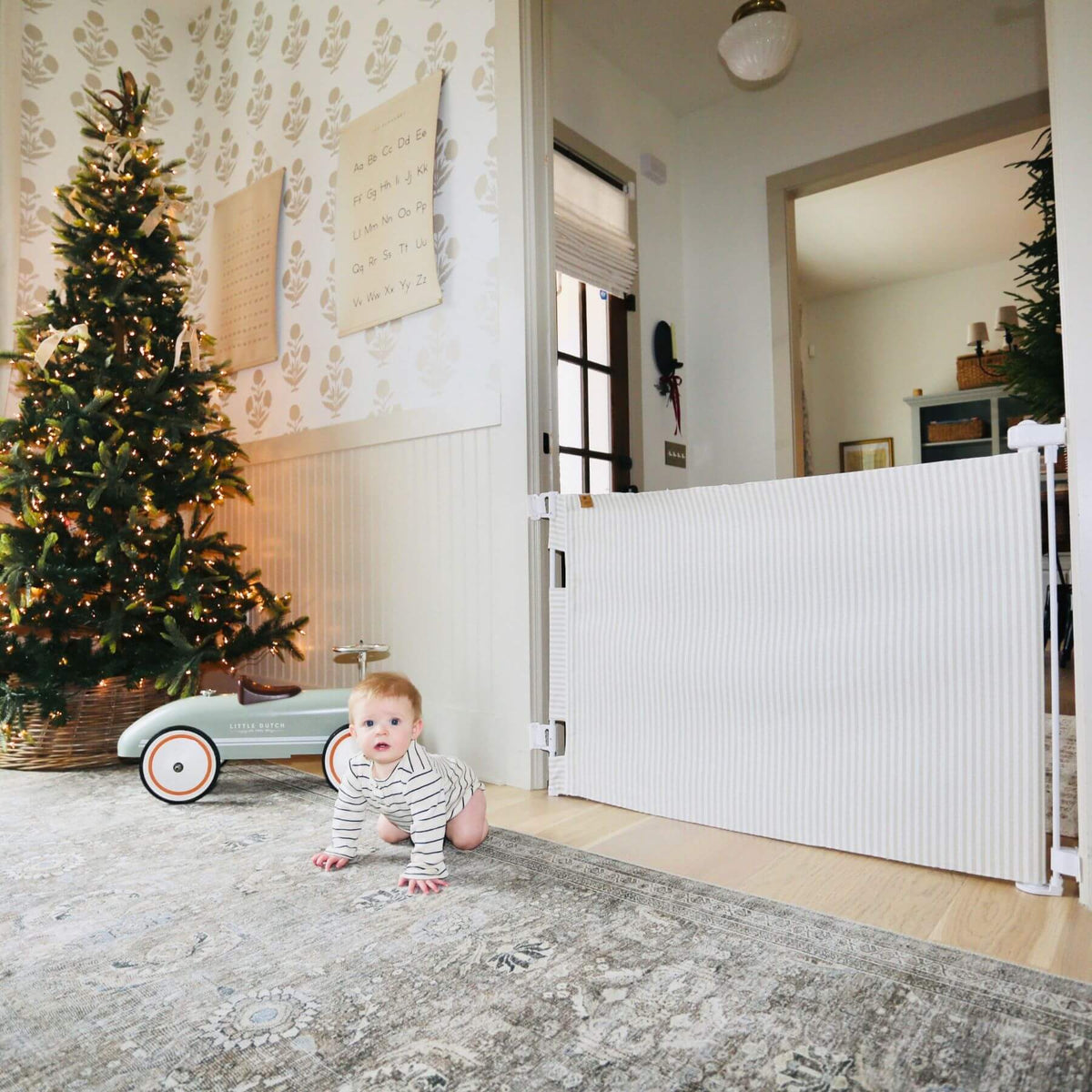 A baby in a striped onesie crawls on a patterned rug in a decorated home interior, with a white ribbed safety gate blocking the doorway and a Christmas tree and toy car nearby.