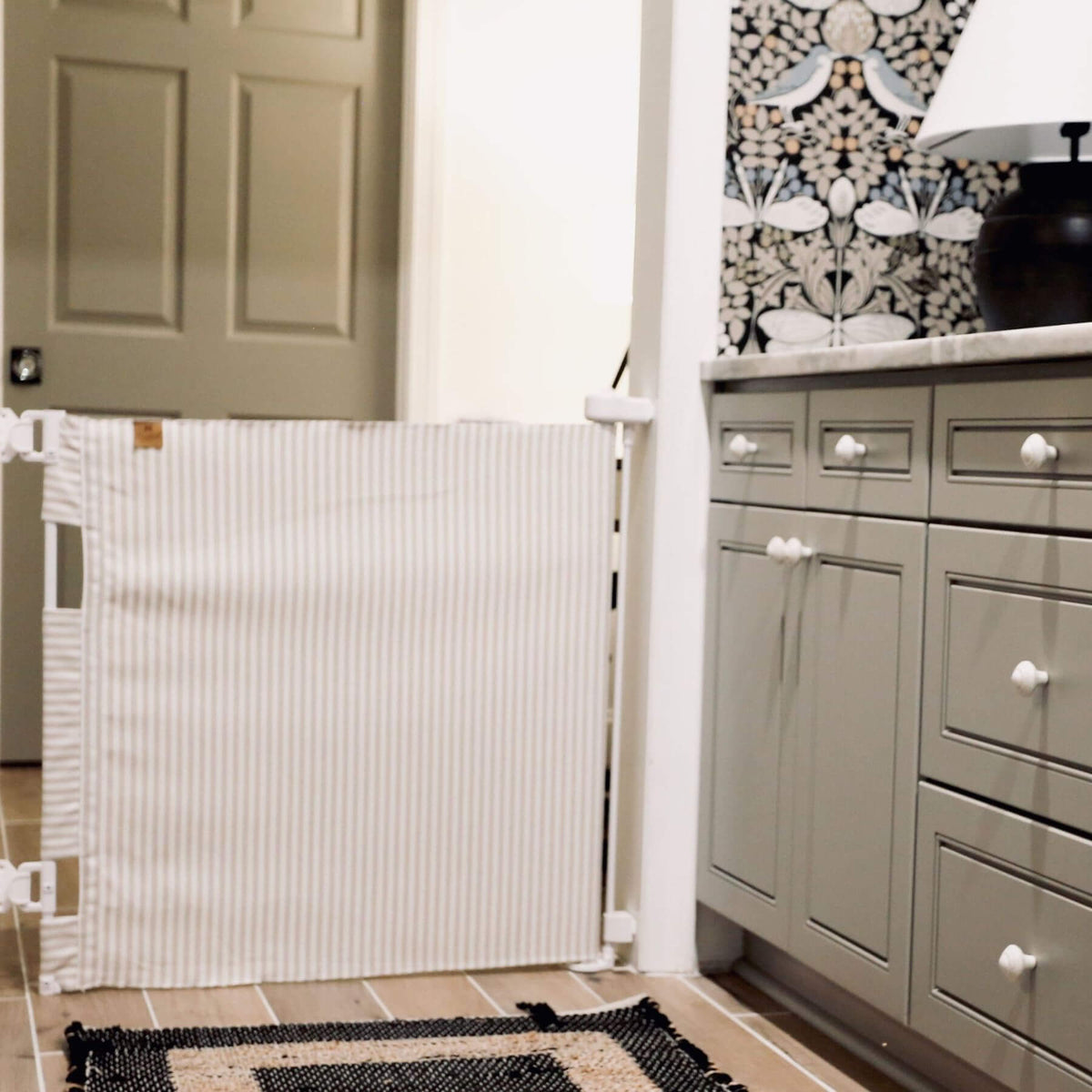 A white fabric baby safety gate with thin vertical stripes, mounted on a white metal frame in a kitchen doorway.
