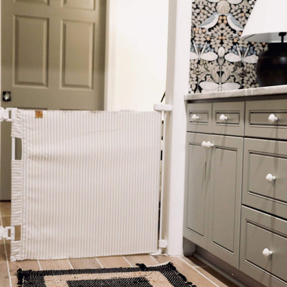 A white fabric baby safety gate with thin vertical stripes, mounted on a white metal frame in a kitchen doorway.