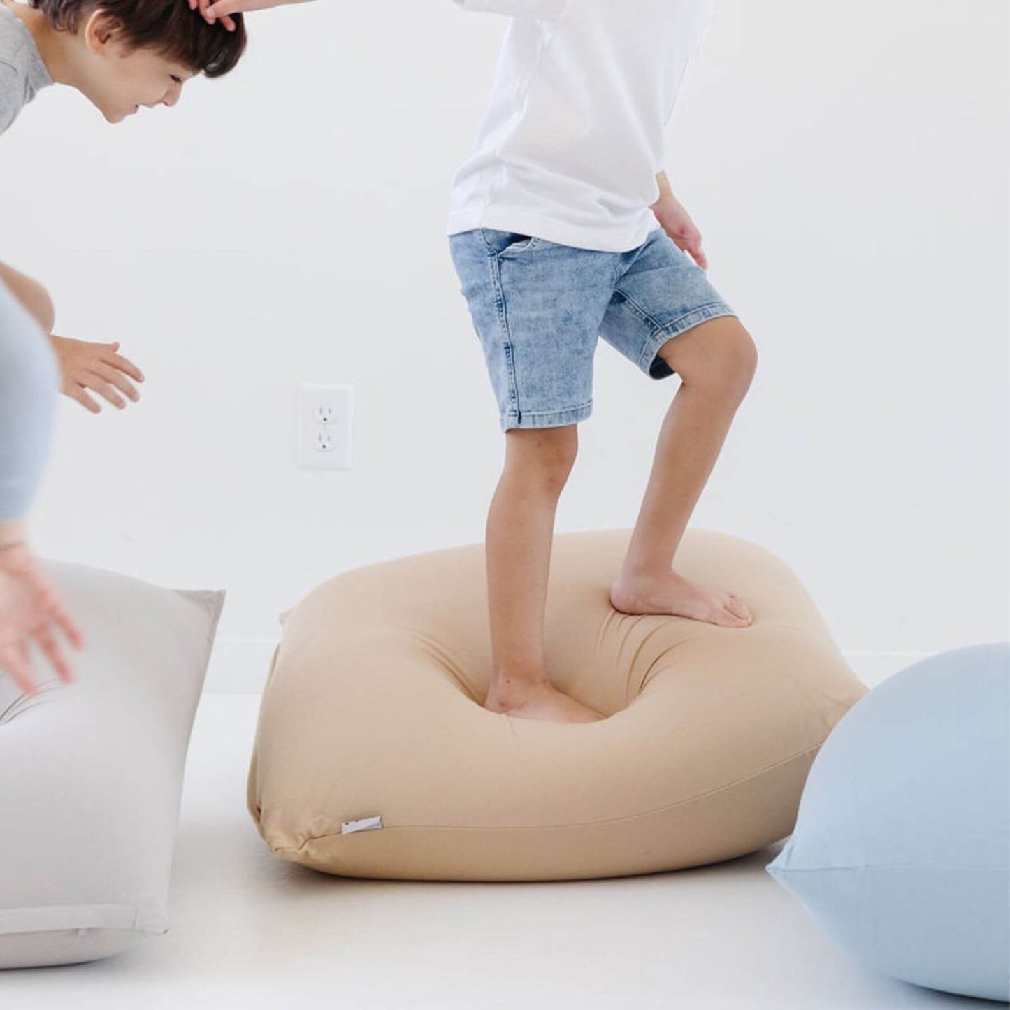 A child stands barefoot on Toki Kids Sandcastle Bean Bag. Another child is partially visible on the left touching a light gray bean bag, and a light blue bean bag is visible on the right.
