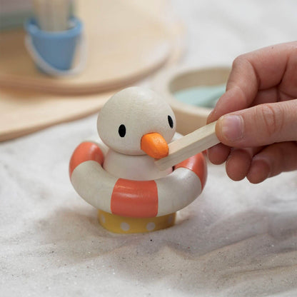 Wooden duck toy with an orange life preserver and a hand holding a stick on a sandy surface.