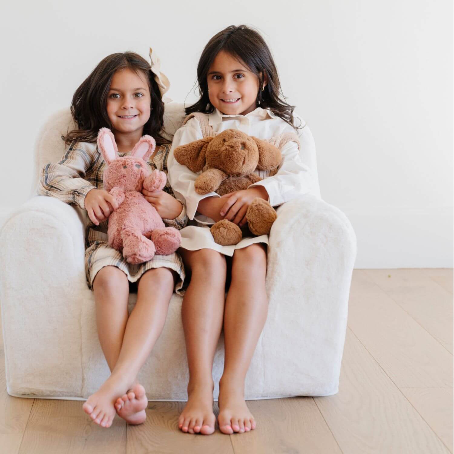 Two young girls sitting on a white chair holding stuffed animals.