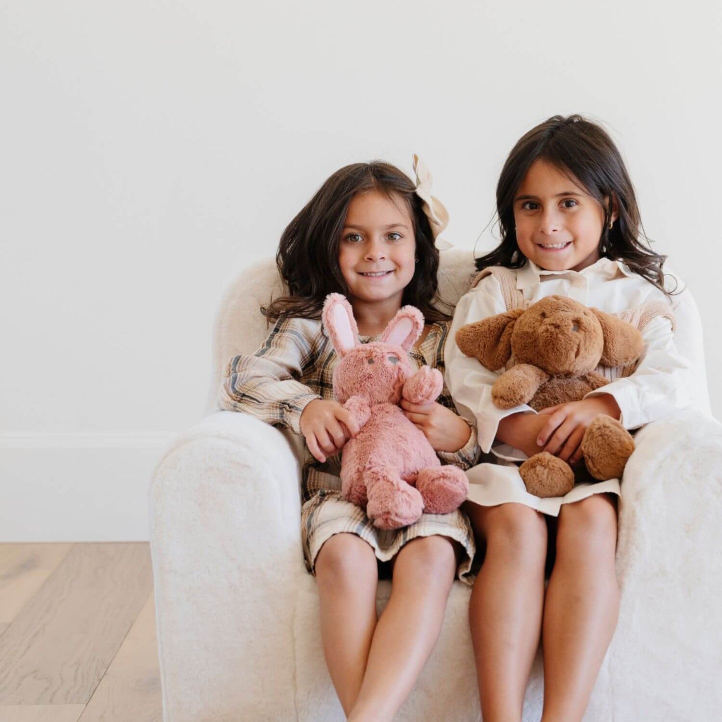 Two young girls sitting on a white chair holding stuffed animals.
