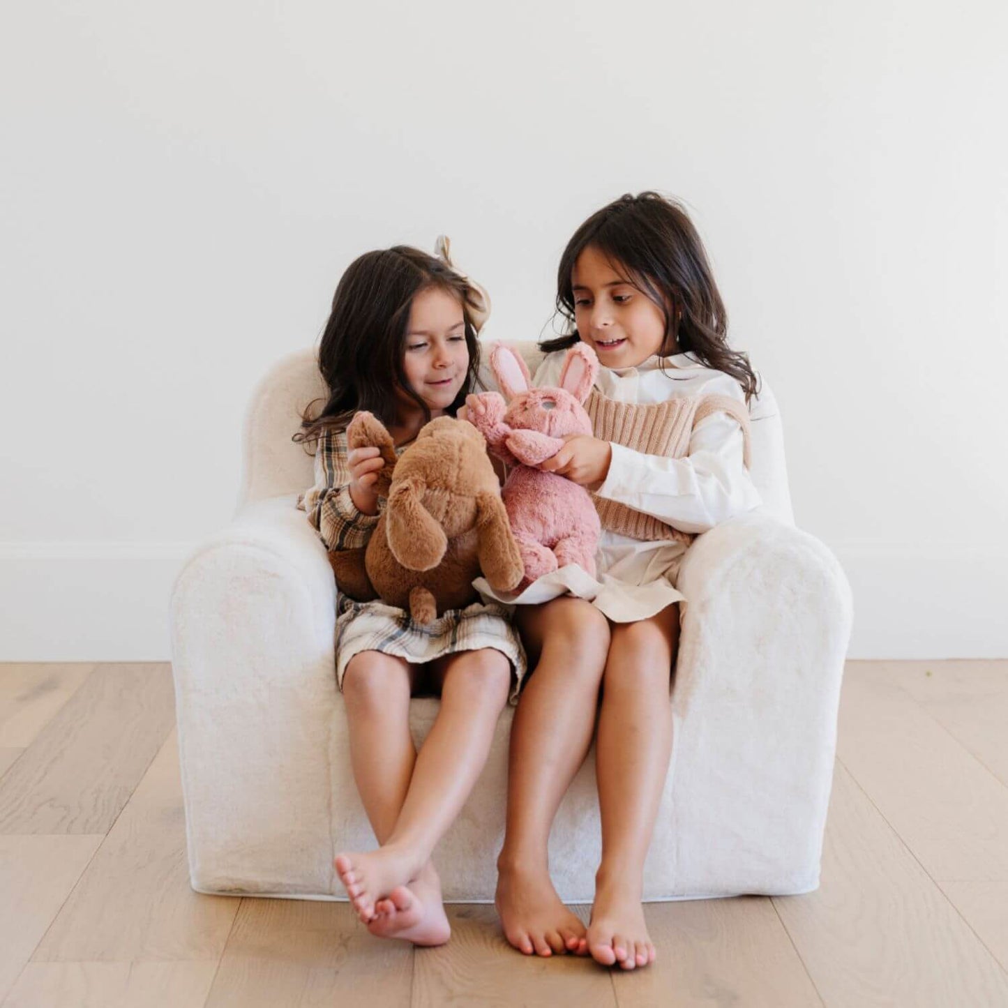 Two children sitting on a white chair holding stuffed animals in a bright room.