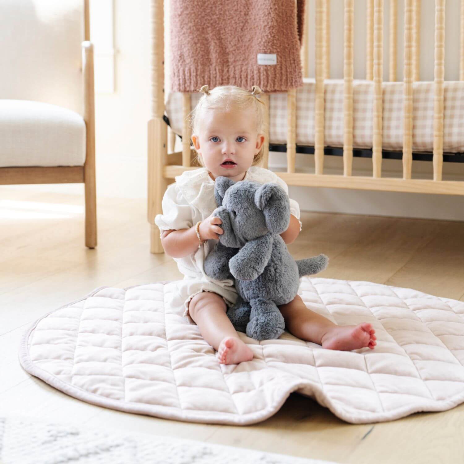 Child sitting on a quilted mat holding a plush toy in a nursery.
