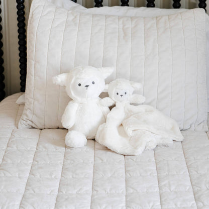 Two Saranoni Stuffed Animal lambs on a striped bedspread with a pillow in the background.