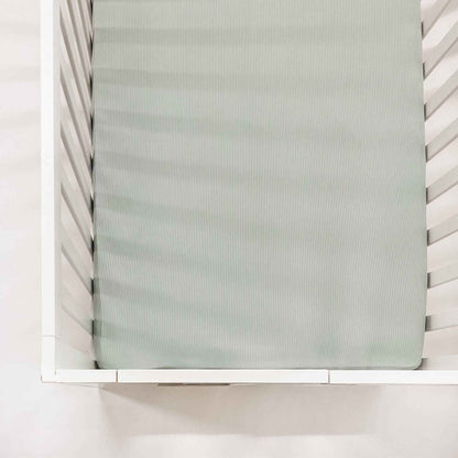 White crib with a mattress covered with Little Mae & Co Seafoam Ribbed Crib Sheet