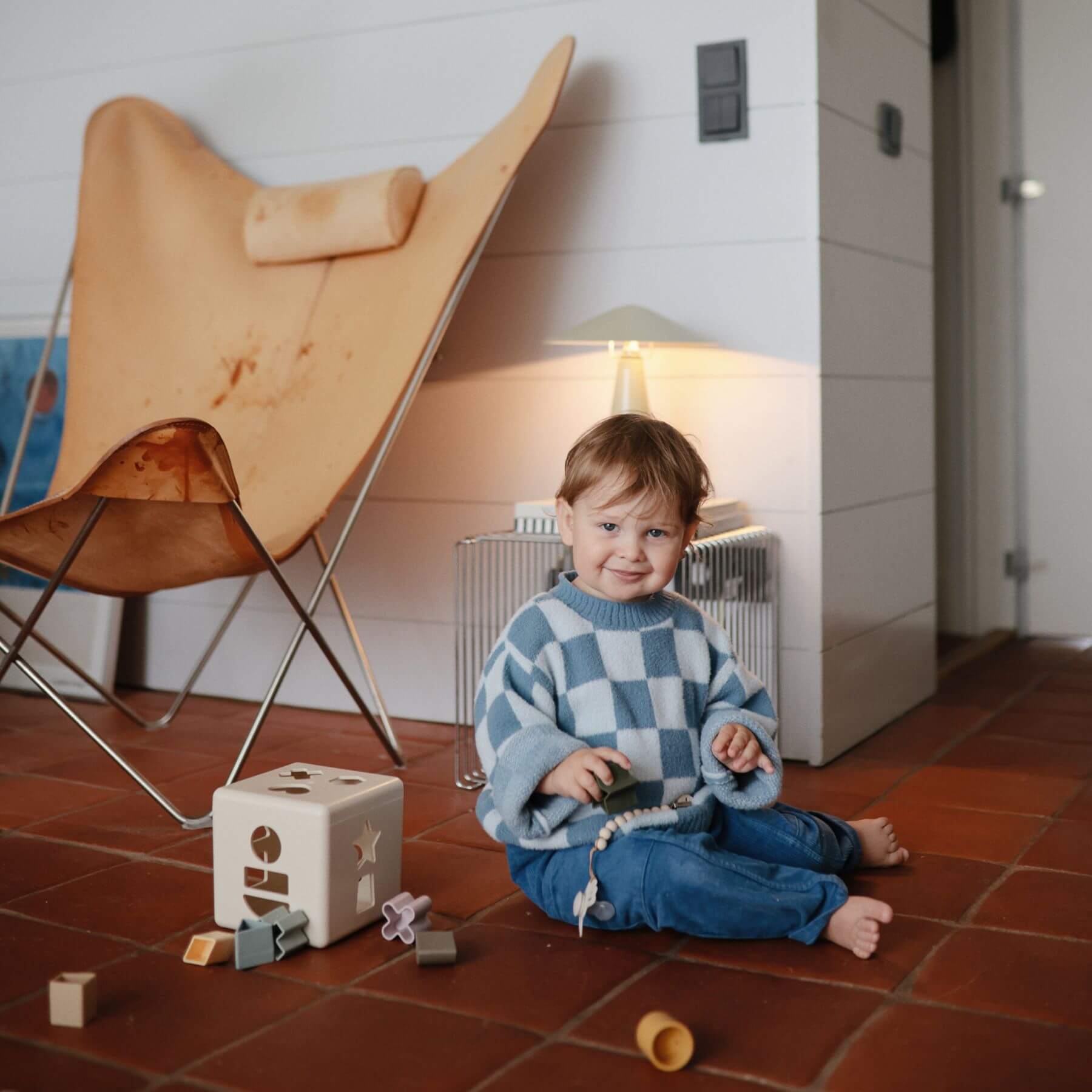 Child sitting on a tiled floor with Mushie Shape Sorting Box, wearing a blue checkered sweater and jeans, in a room with a wooden chair and lamp.