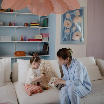 Woman and child sitting on a couch with Mushie Shape Sorting Box, in a room with shelves and artwork.