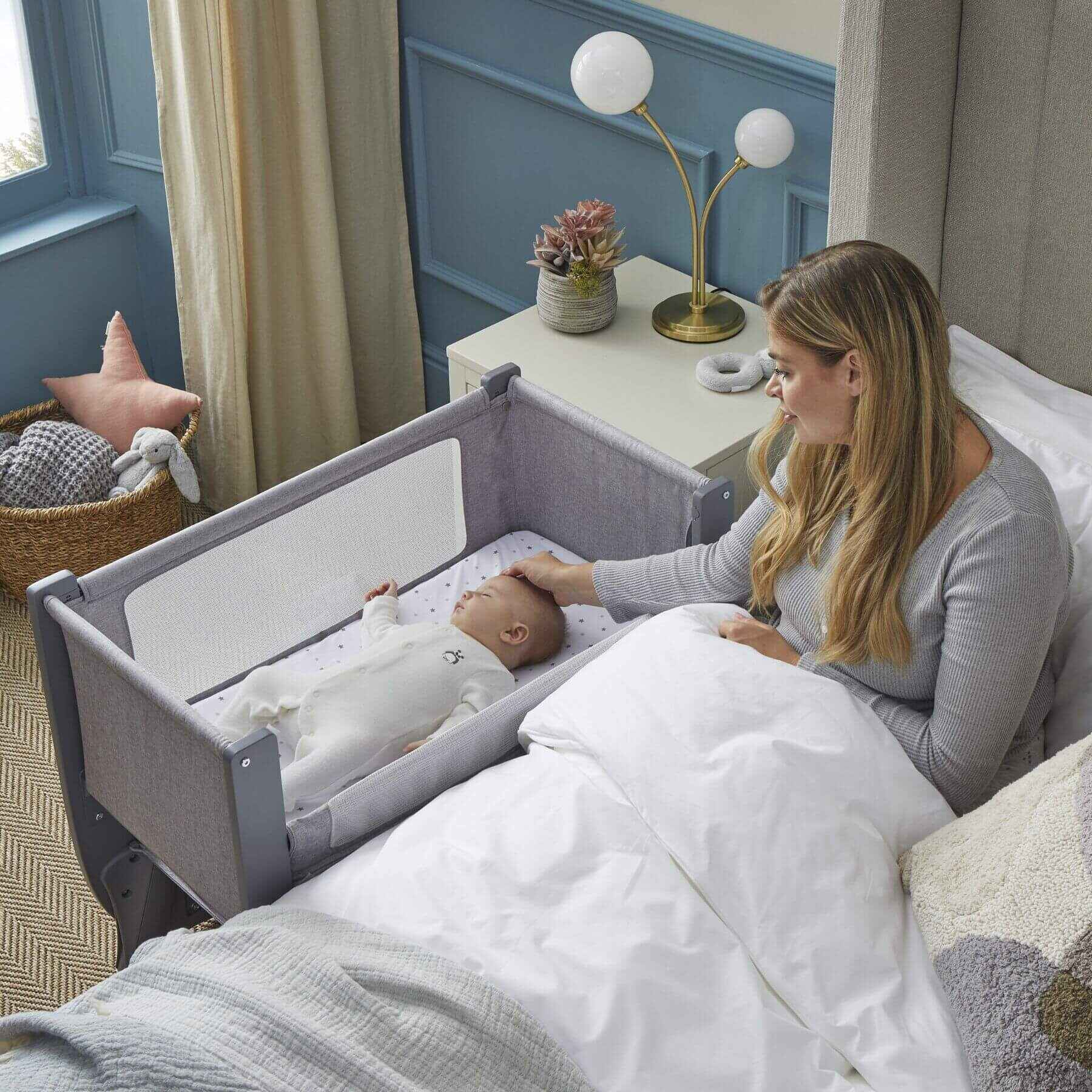 Woman tending to a baby in a crib next to her bed in a bedroom.