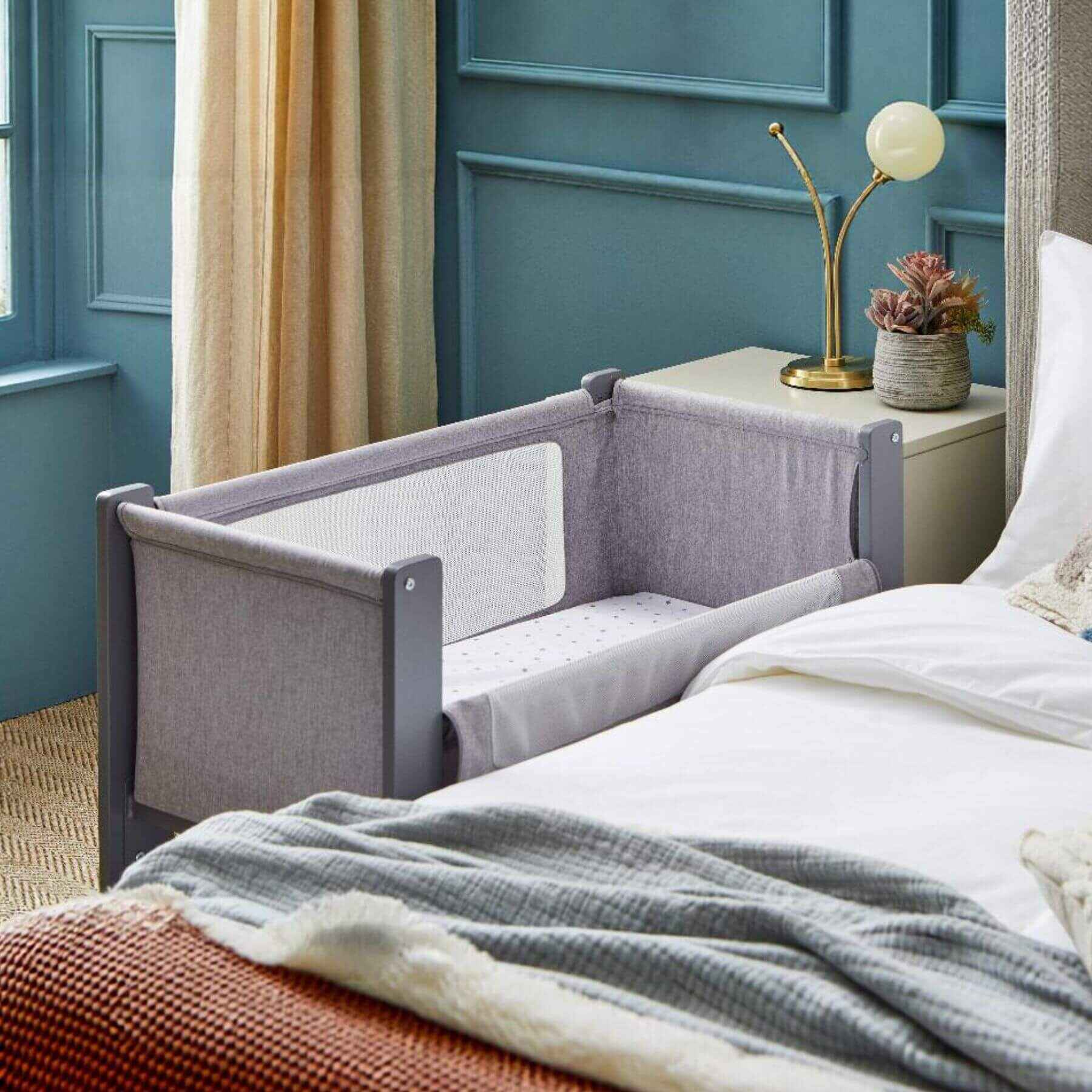 Gray crib attached to a bed in a bedroom with blue walls and a lamp.