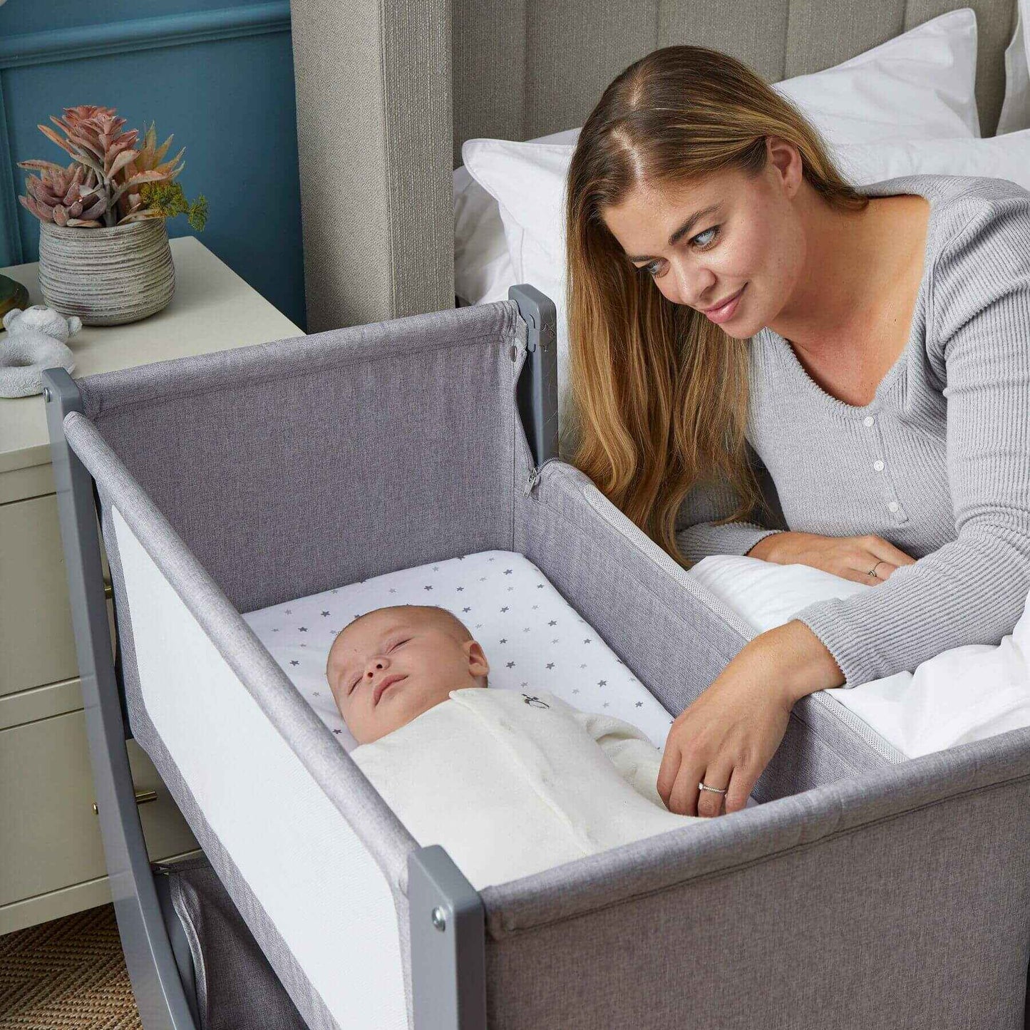 Woman sitting next to a baby in a gray crib within a bedroom setting.