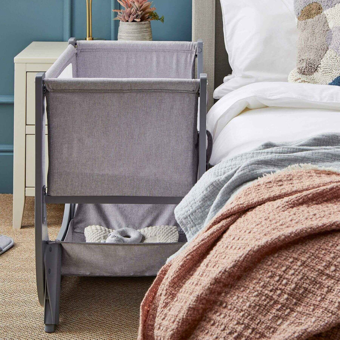 Gray bedside crib next to a bed with pink blanket in a bedroom setting.