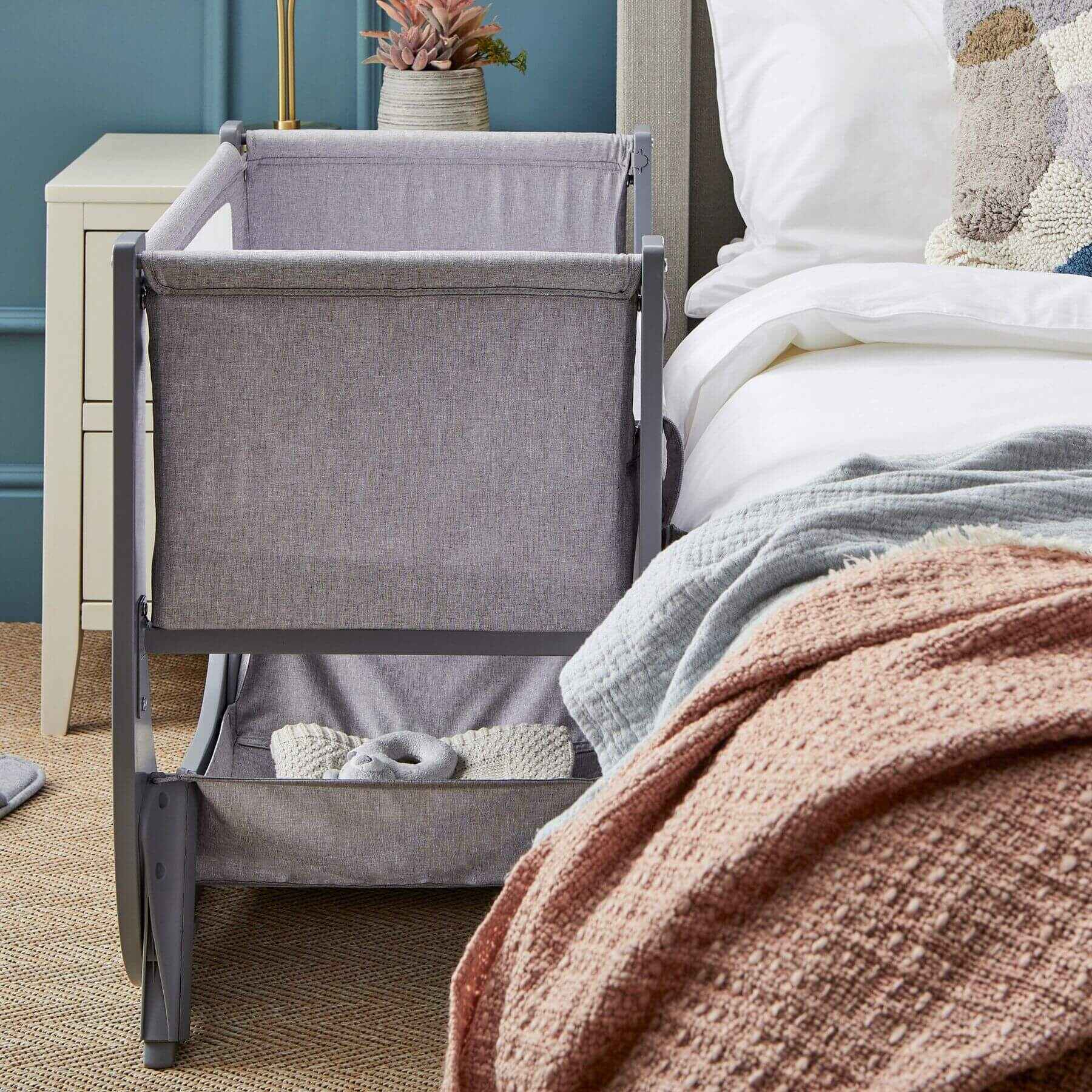 Gray bedside crib next to a bed with pink blanket in a bedroom setting.