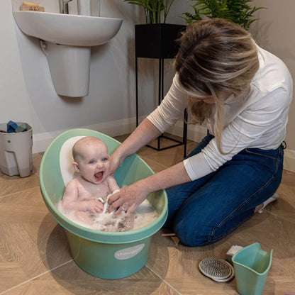 Woman bathing a baby in a green basin in a bathroom.