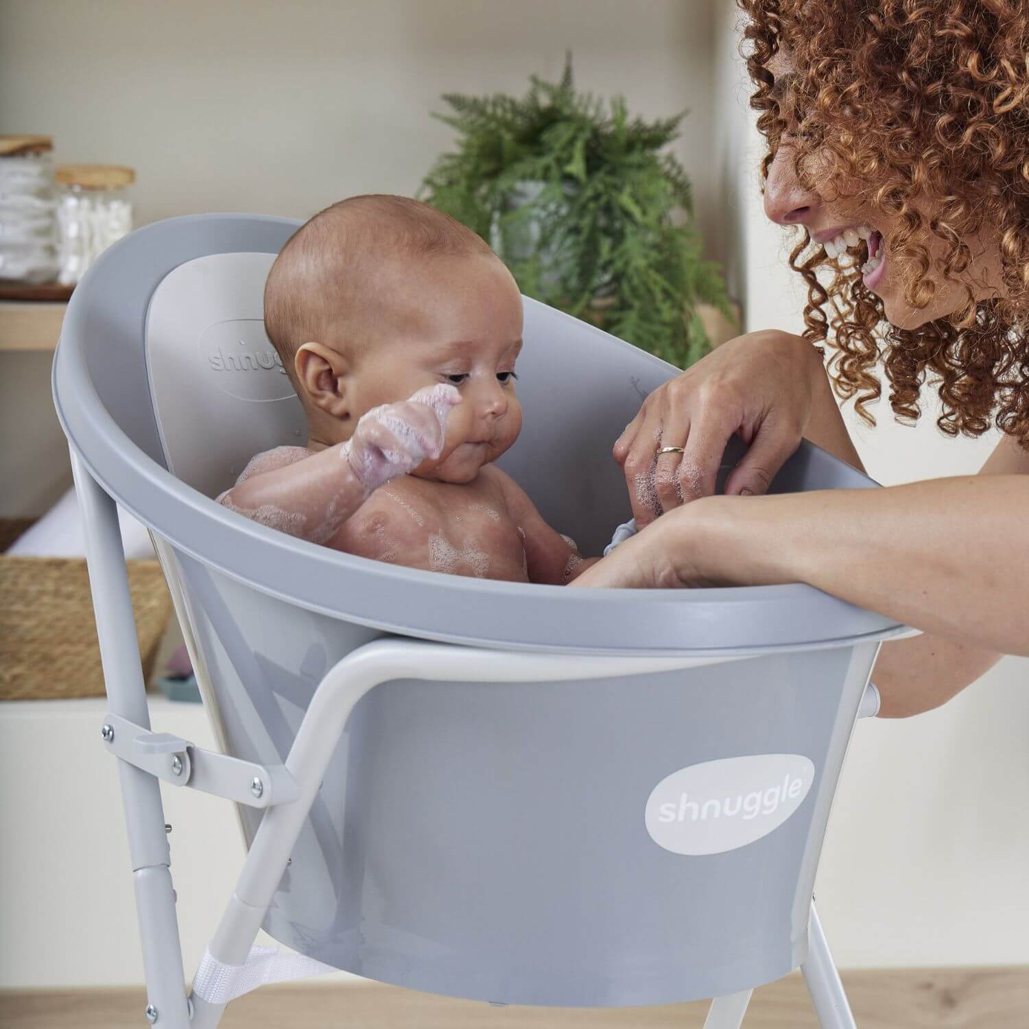 Baby in a gray Snuggles baby bath seat with a woman beside it.