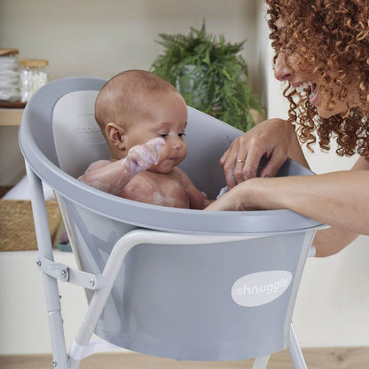 Baby in a gray Snuggles baby bath seat with a woman beside it.