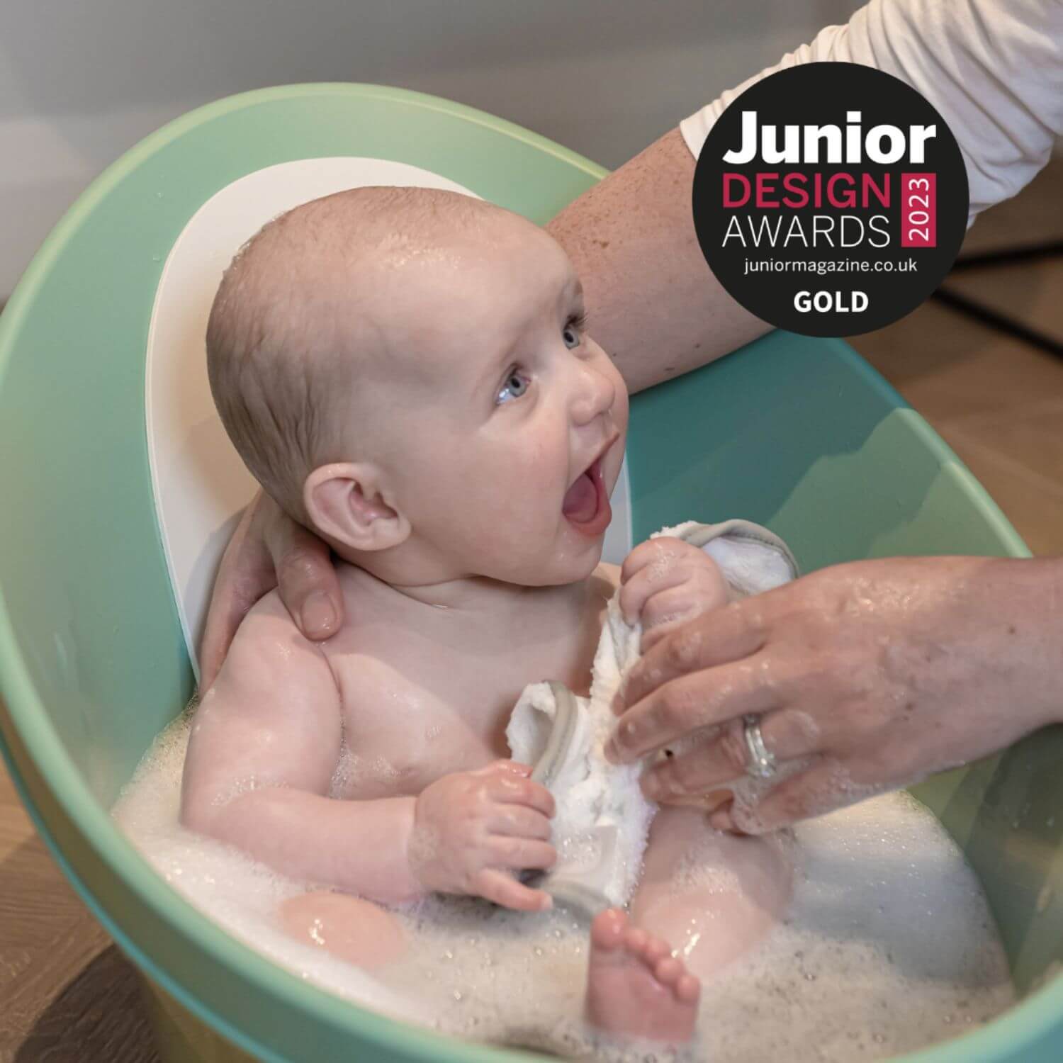 Baby being bathed in a green basin with a person, featuring the Junior Design Awards 2023 logo.