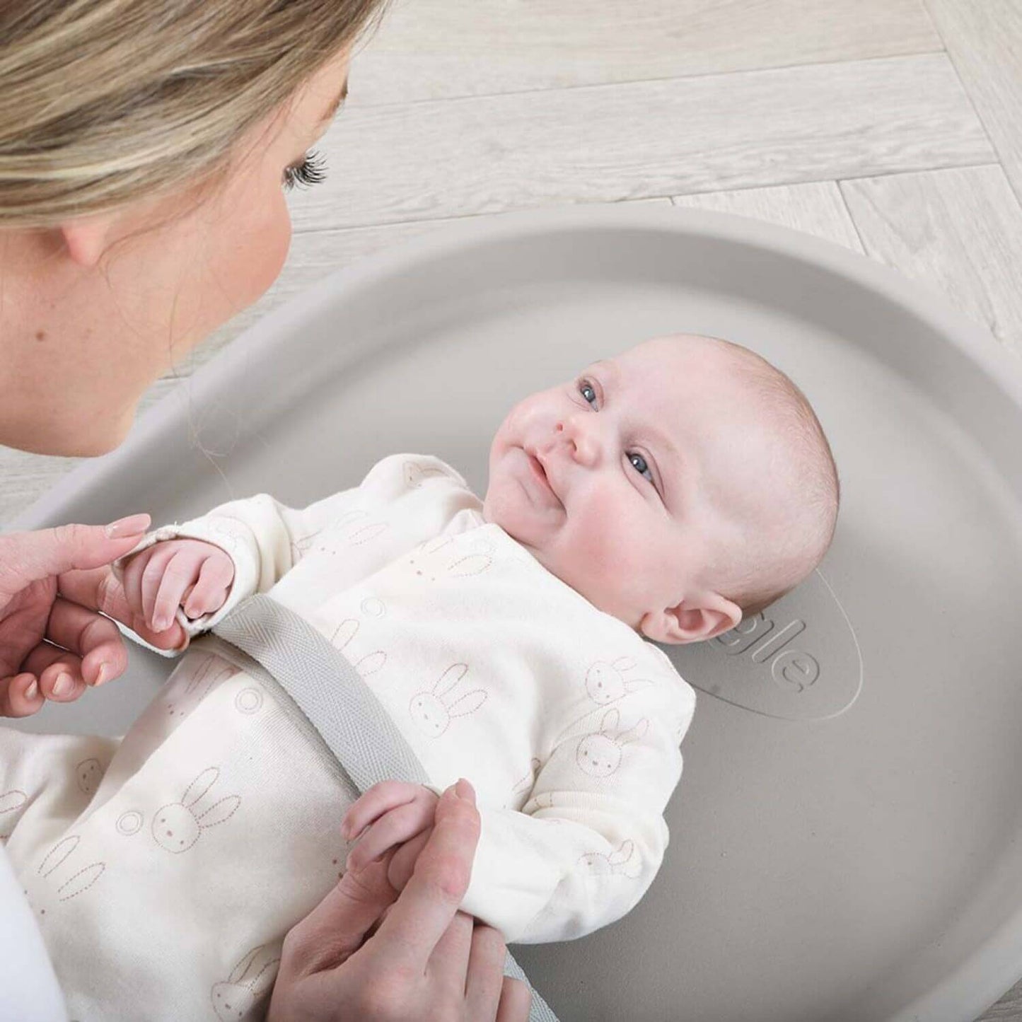 Baby lying in a gray changing tray with a woman adjusting a strap, on a light wooden floor.