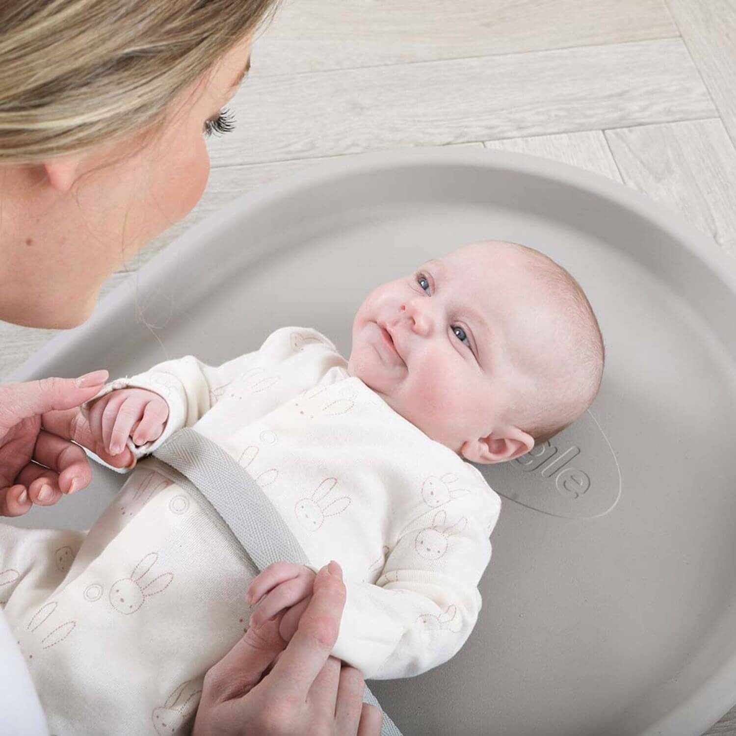 Baby lying in a gray changing tray with a woman adjusting a strap, on a light wooden floor.