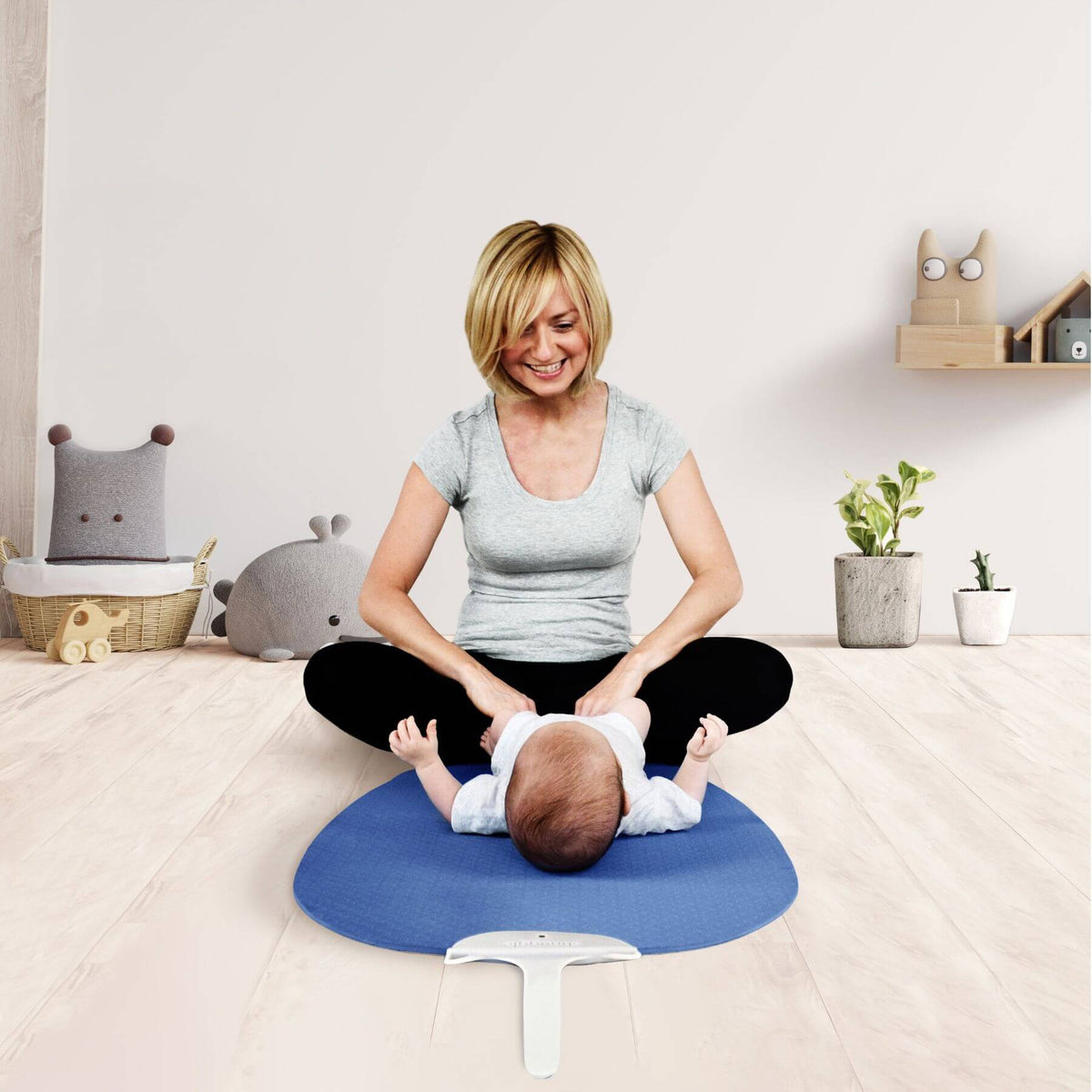A woman sits cross-legged on a light wood floor in a nursery, using a blue baby changing pad with a baby lying on their stomach.