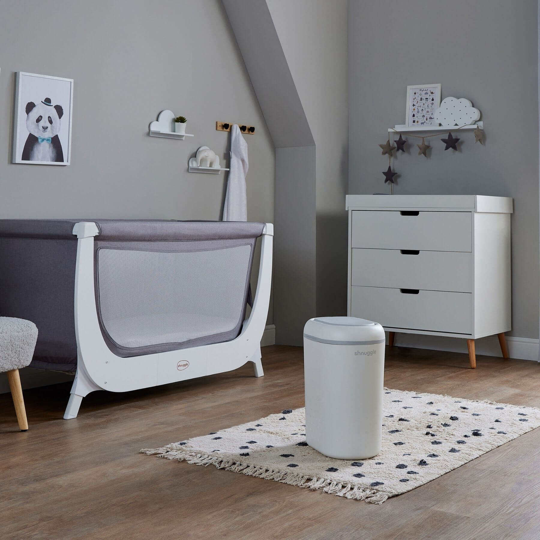 A modern baby nursery room featuring a white and gray convertible crib, a white three-drawer dresser with light wood legs, Shnuggle Eco-Touch Nappy Bin Diaper Pail - White, and a patterned rug on a wood floor.