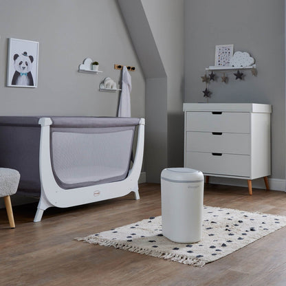 A modern baby nursery room featuring a white and gray convertible crib, a white three-drawer dresser with light wood legs, Shnuggle Eco-Touch Nappy Bin Diaper Pail - White, and a patterned rug on a wood floor.