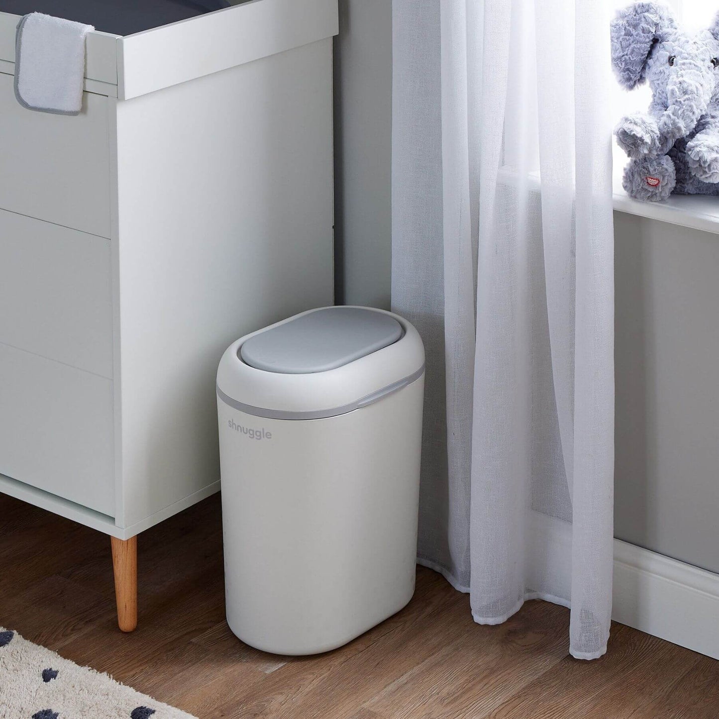 Shnuggle Eco-Touch Nappy Bin Diaper Pail - White, placed next to a white cabinet in a room with wood flooring, a sheer curtain, and a plush toy on a windowsill.