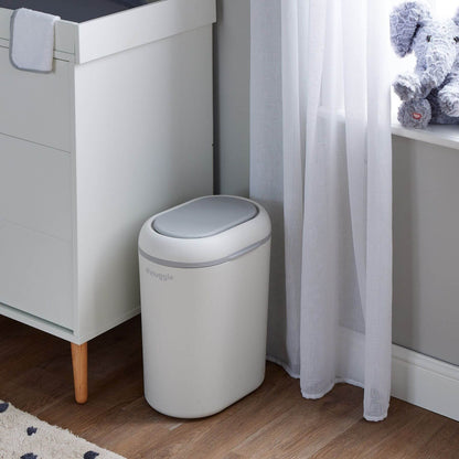 Shnuggle Eco-Touch Nappy Bin Diaper Pail - White, placed next to a white cabinet in a room with wood flooring, a sheer curtain, and a plush toy on a windowsill.