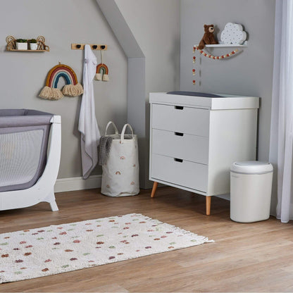 A white three-drawer dresser with light wood legs in a styled nursery setting, next to a white changing table, a gray laundry hamper, and Shnuggle Eco-Touch Nappy Bin Diaper Pail - White on a light wood floor.