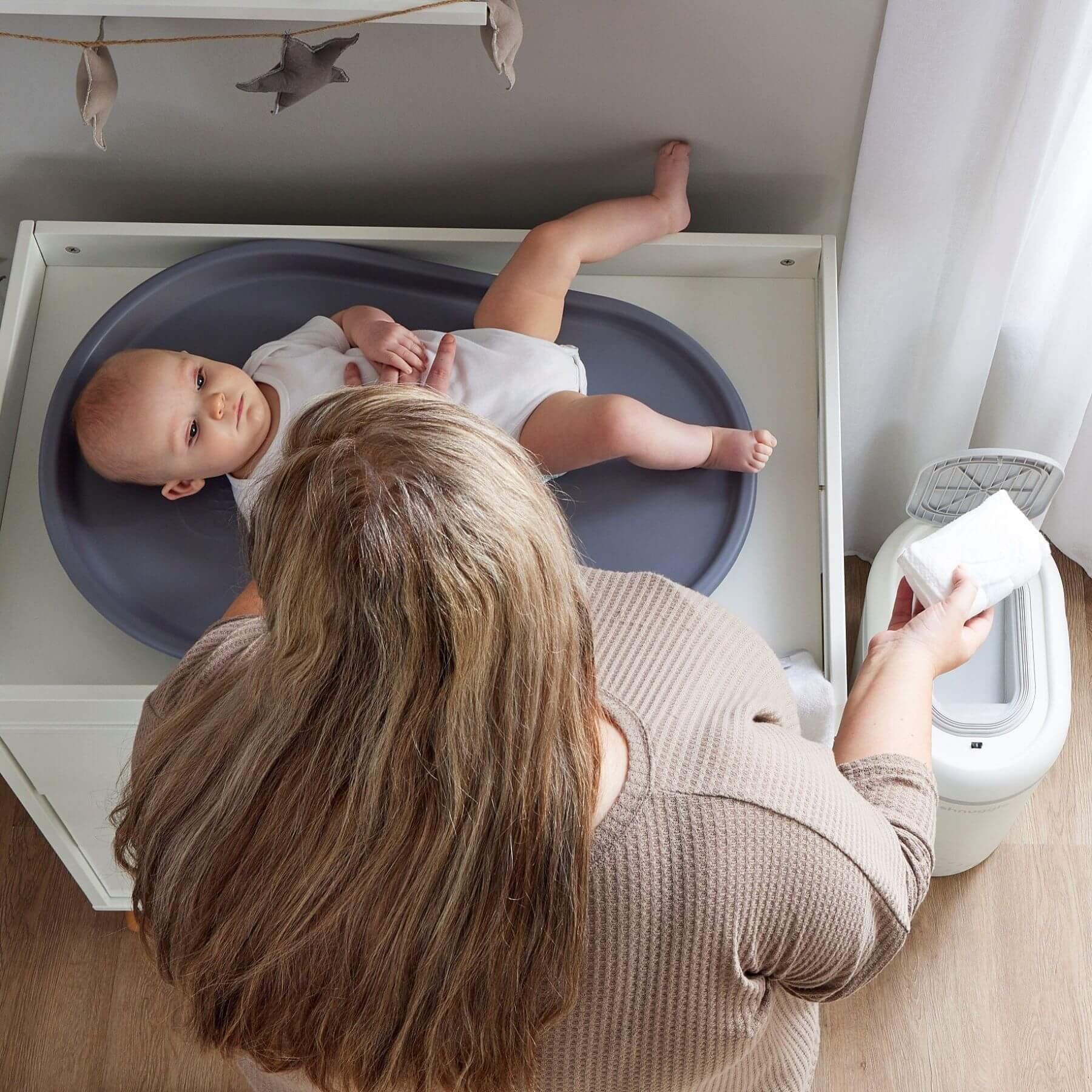 A baby changing scene with a baby lying on a gray changing tray on a white changing table, a woman holding wipes in the foreground, and Shnuggle Eco-Touch Nappy Bin Diaper Pail - White