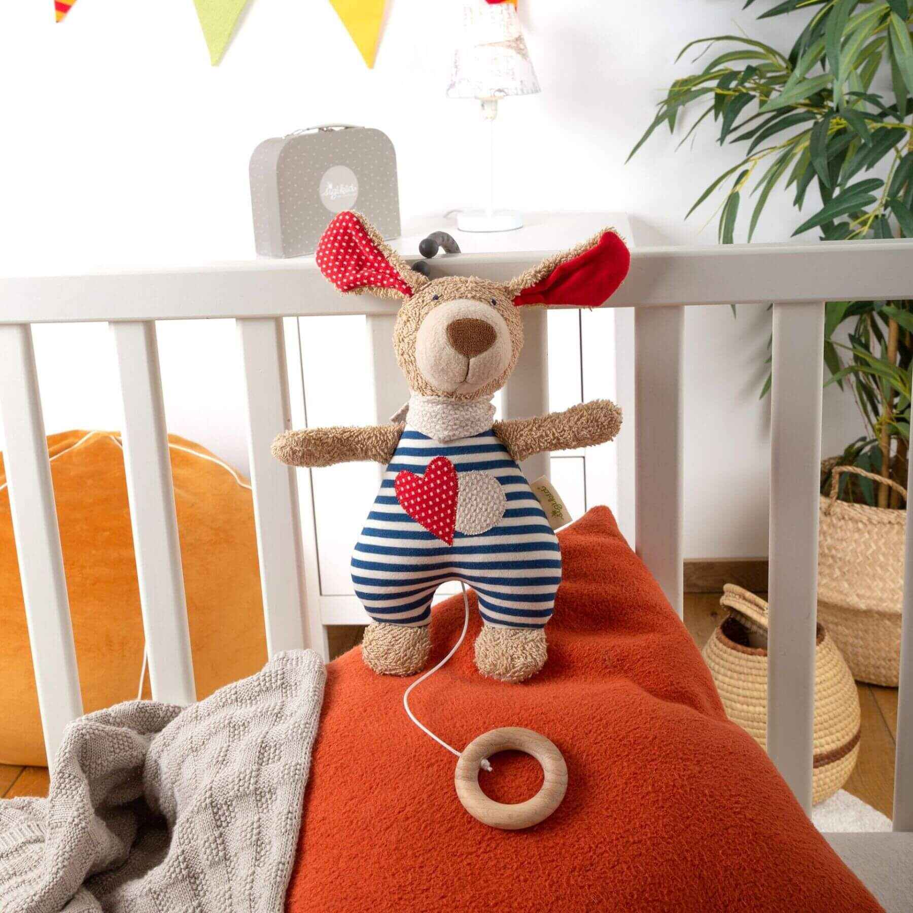 sigikid Organic Dog Musical Toy in a striped outfit on an orange cushion next to a crib.