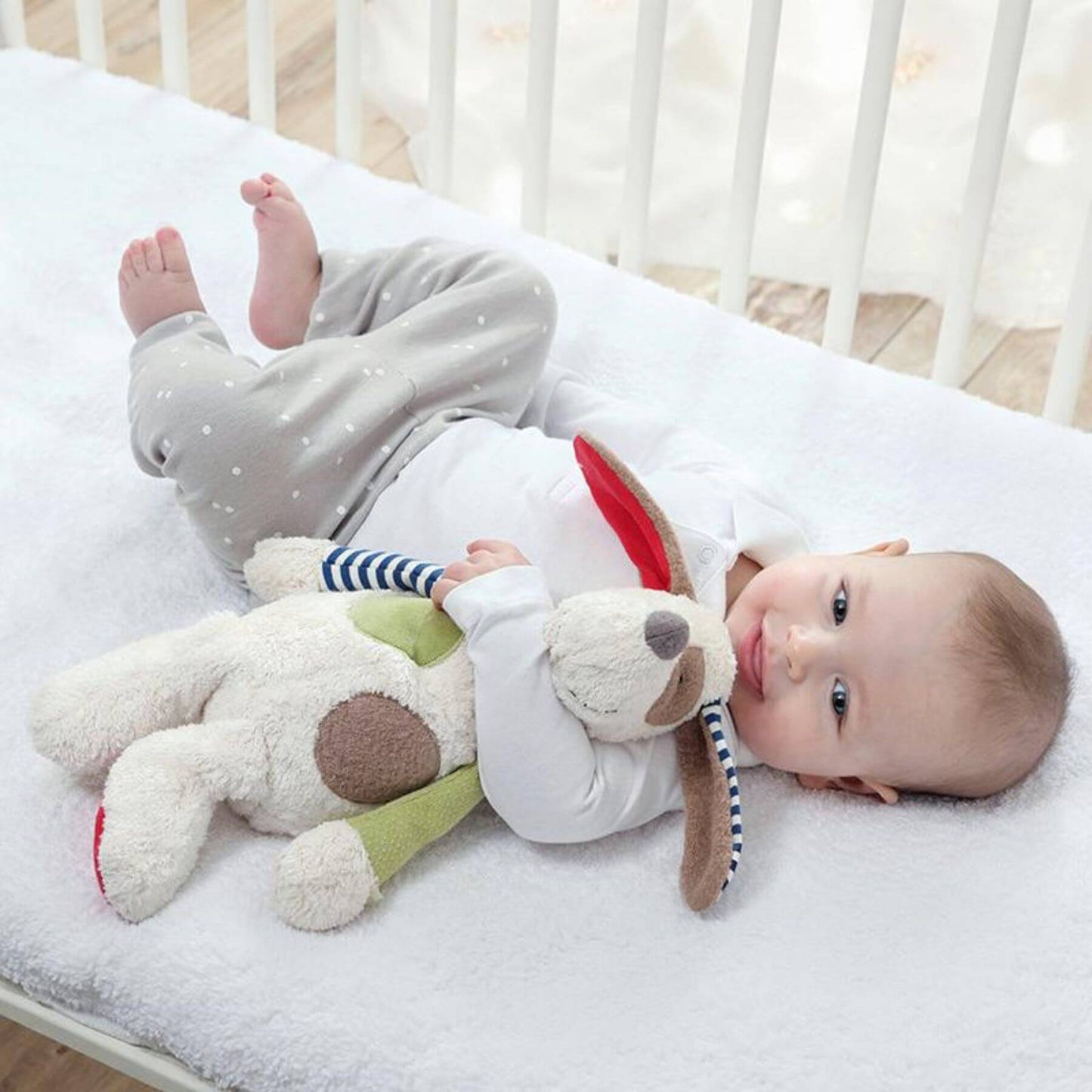 Baby lying on a bed with sigikid Organic Dog Plush Toy, wearing a white shirt and gray pants.