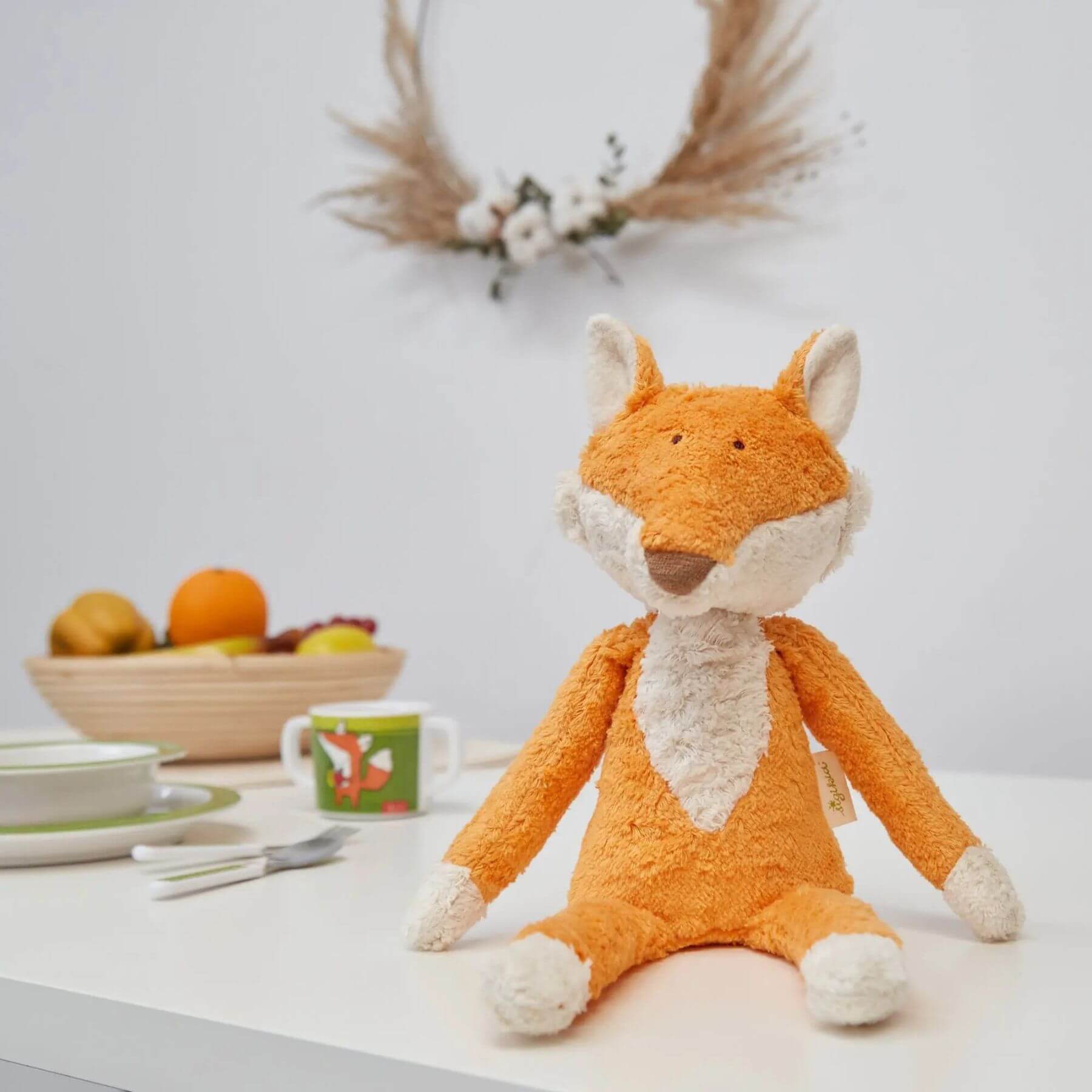 sigikid Organic Plush Fox on a table with a bowl of fruit and decorative items in the background.